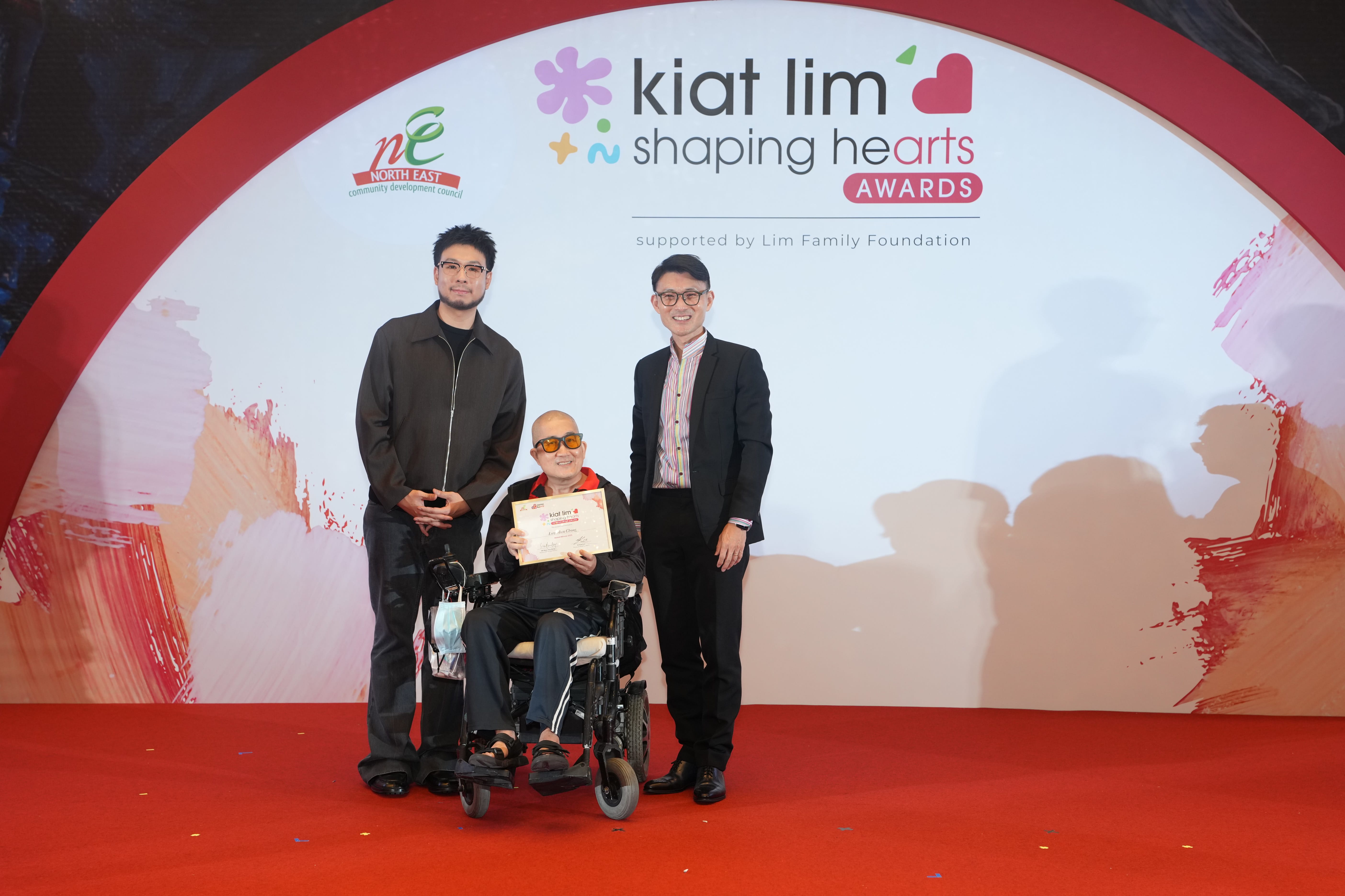 Award recipient standing on stage with the presenter, holding their certificate and posing for a group photo during the award ceremony, with a backdrop reading ‘Kiat Lim-Shaping Hearts Awards'.